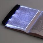 A transparent LED reading light panel placed over a paperback book page in a dark room.
